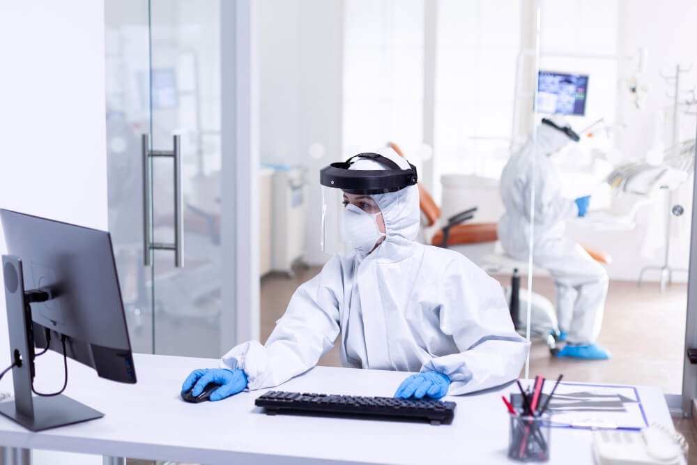 nurse-using-computer-during-covid-19-wearing-ppe-suit-as-safety-precaution-medicine-team-wearing-protection-gear-against-coronavirus-pandemic-dental-reception-as-safety-precaution